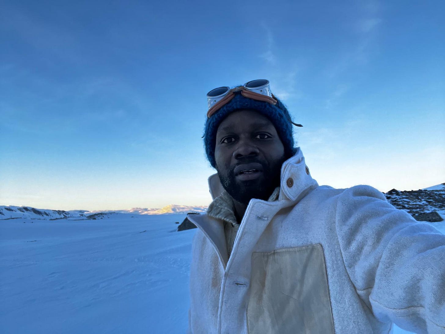 Dwayne Fields standing in a snowy Arctic landscape at sunrise, wearing a thick white expedition coat and blue hat with goggles, under a wide clear sky.