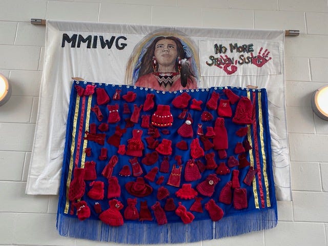 An installation featuring a large fabric banner hung on a white brick wall. The top portion is a white cloth with the text “MMIWG” on the left and “No More Stolen Sisters” on the right, accompanied by red handprints. Below it hangs a vibrant blue panel decorated with numerous small, handmade red garments and accessories—such as tiny dresses, hats, mittens, and other knit or crocheted items—attached in a grid-like arrangement. Yellow and blue ribbon accents frame the sides of the blue panel, and blue fringe hangs along the bottom edge. Two circular wall lights sit on either side of the installation. An installation featuring a large fabric banner hung on a white brick wall. The top portion is a white cloth with the text “MMIWG” on the left and “No More Stolen Sisters” on the right, accompanied by red handprints. Below it hangs a vibrant blue panel decorated with numerous small, handmade red garments and accessories—such as tiny dresses, hats, mittens, and other knit or crocheted items—attached in a grid-like arrangement. Yellow and blue ribbon accents frame the sides of the blue panel, and blue fringe hangs along the bottom edge. Two circular wall lights sit on either side of the installation.