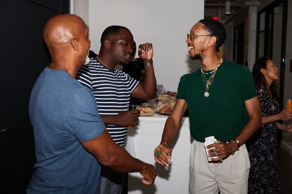 Three men standing and talking at a social event, with a table of food behind them. 