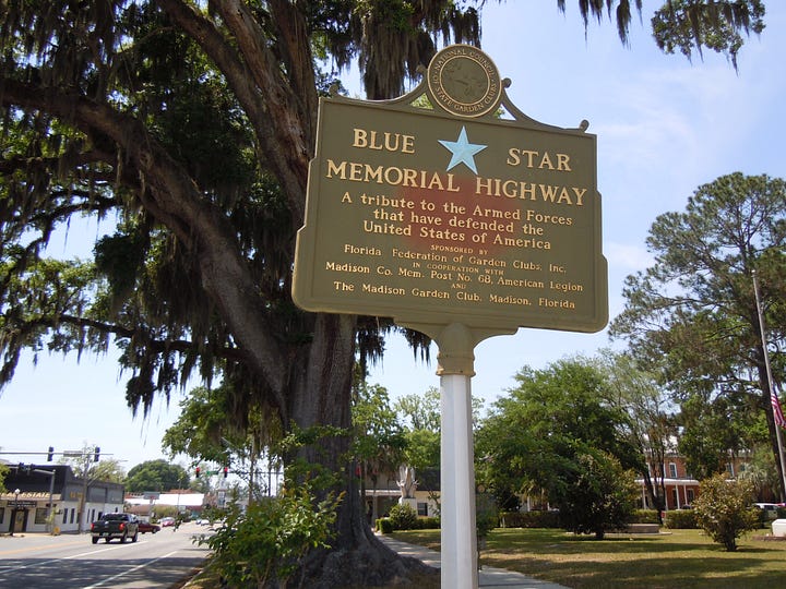 Portrait of a young Edward Hood in a dark military dress uniform with U.S. flags in the background and Blue Star Memorial Highway sign honoring the U.S. Armed Forces beneath a large moss-draped tree by a roadside.