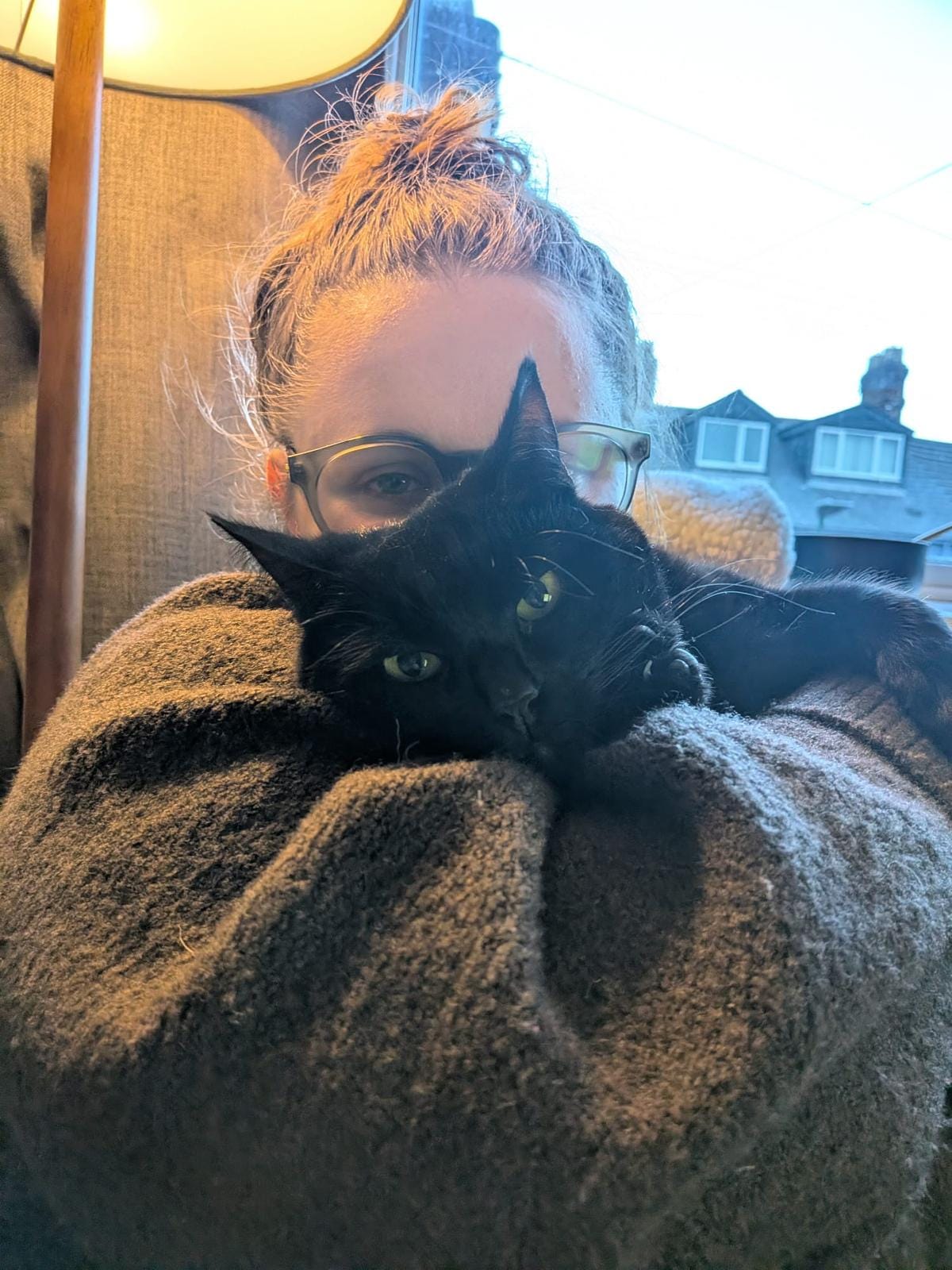 Lucy Rose cuddles her black cat Figgy in a cosy window-lit selfie, bundled in a soft blanket at home on the north-east coast of England, capturing the intimate behind-the-scenes mood of her Tarot DMs interview about writing, creativity, and her debut novel The Lamb.