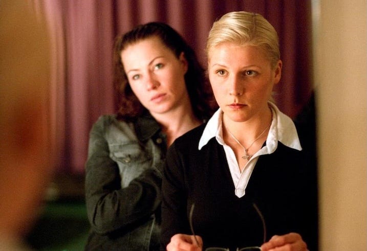 Two women stand in front of a mirror. The blonde woman in front looks serious, holding her glasses, while the brunette behind her seems to observe quietly. The moment feels tense and introspective.