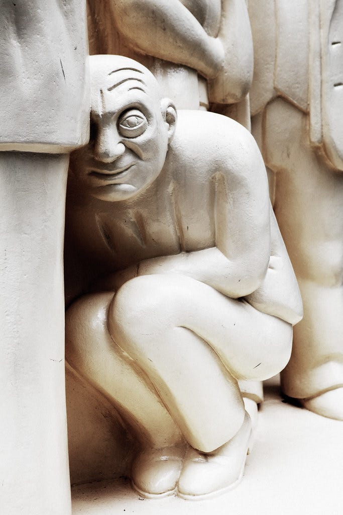 white statue of a man cowering down. he's in a suit and wears a strange smile or a grimace