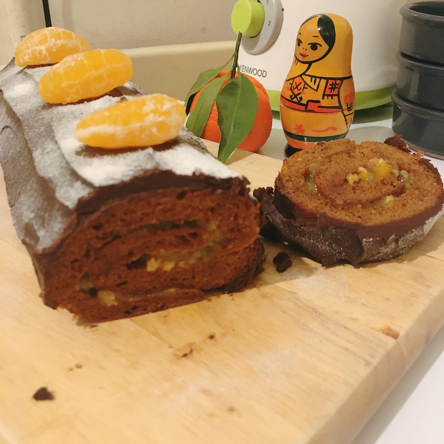On voit la matriochka en bois clair peint, derrière une bûche patissière, près d'une clémentine, devant un mixeur.