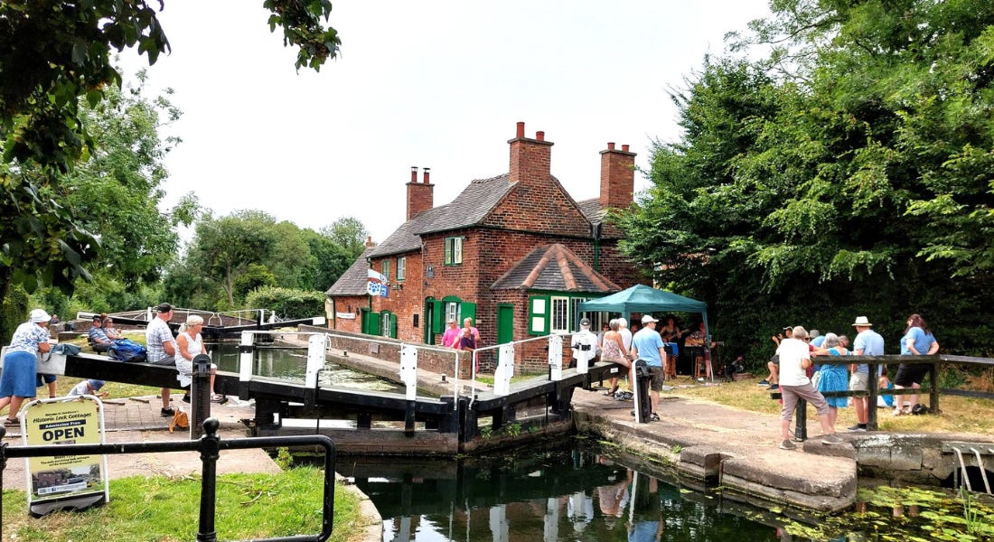A canalside view with people walking over the canal and a brick house with green windows in the distance A canalside view with people walking over the canal and a brick house with green windows in the distance