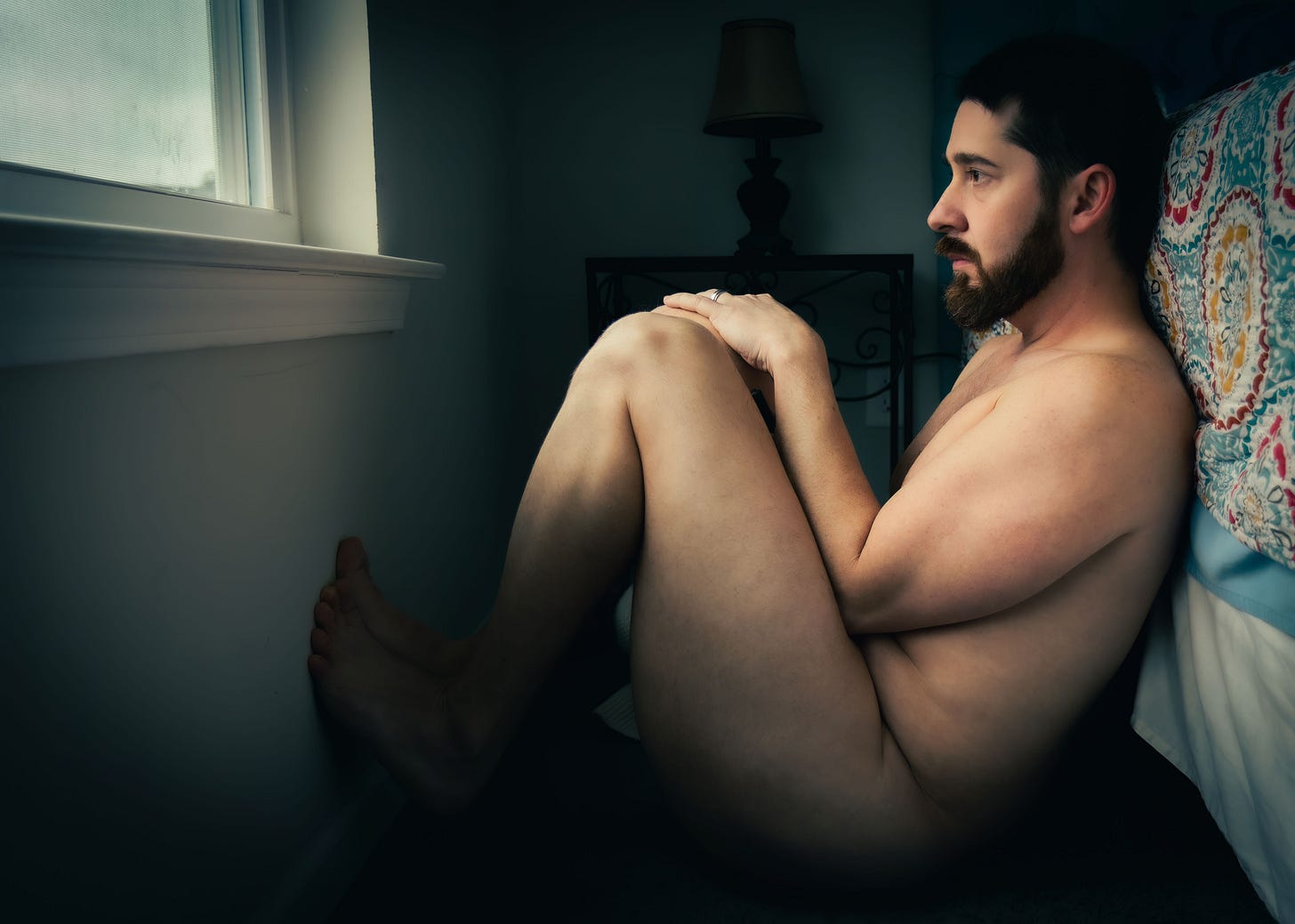 Nude person sitting on the floor beside a bed, knees drawn to their chest, looking toward a window in a dimly lit room.