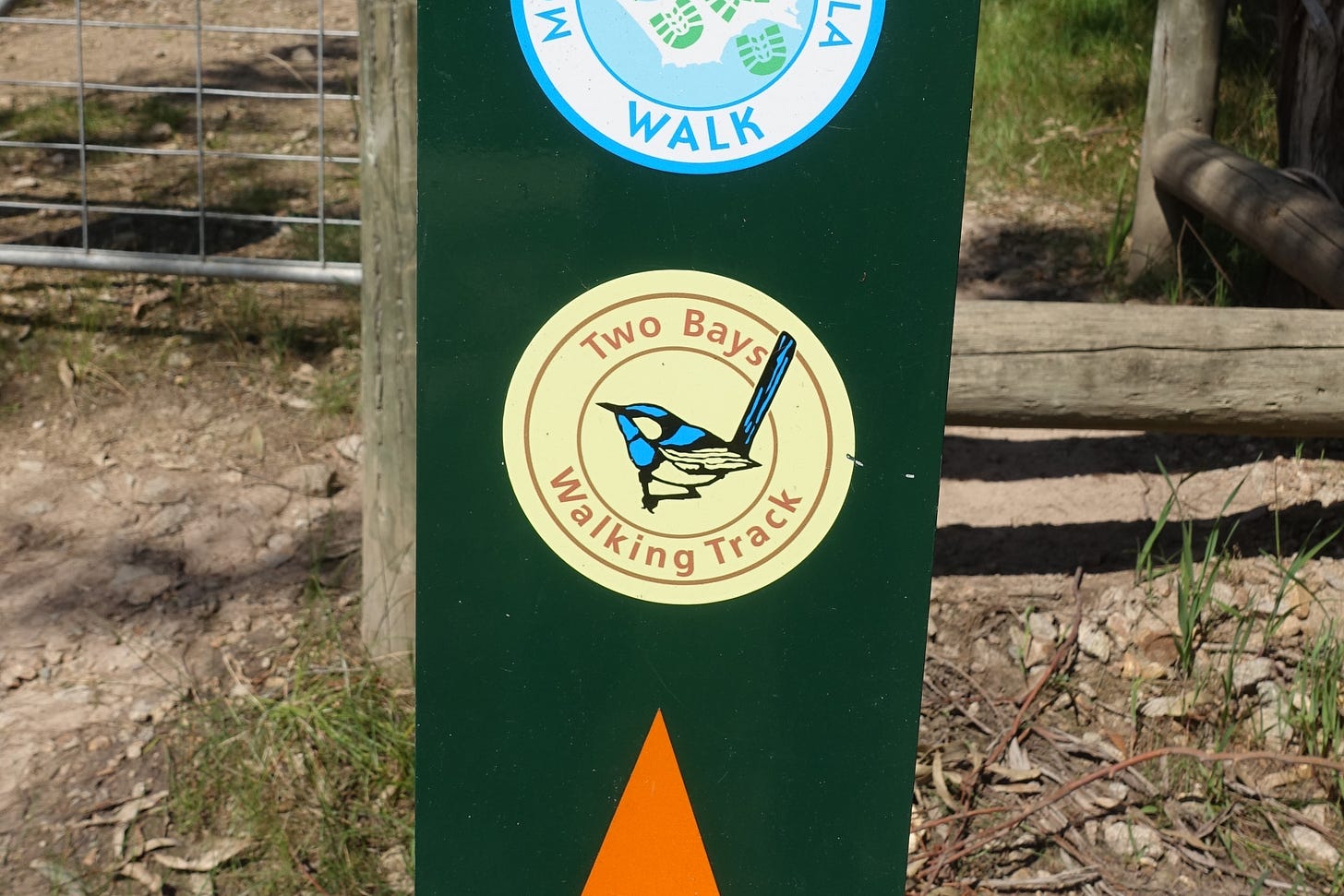 A green trail marker post displaying the 'Two Bays Walking Track' logo featuring a blue fairy wren bird, with an orange directional arrow below