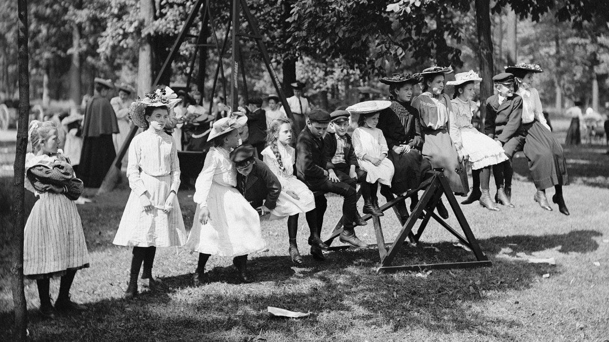 Children in vintage clothing play on a seesaw and gather nearby in a park setting with adults, trees, and classic playgrounds in the background.