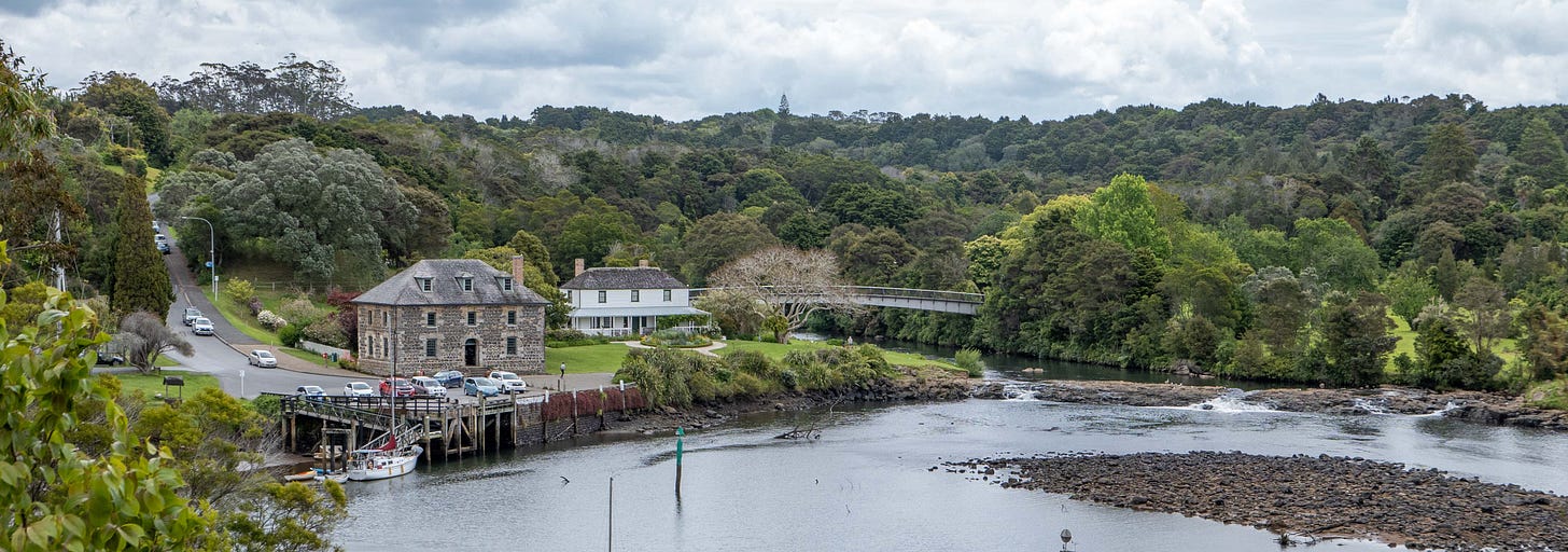 The Stone Store and the Kerikeri Mission House with the river basin in front and a footbridge spanning the river and forested slopes behind.