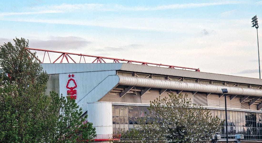 A silver stadium, Nottingham Forest's City Ground, with a tree in front of it and a blue sky above