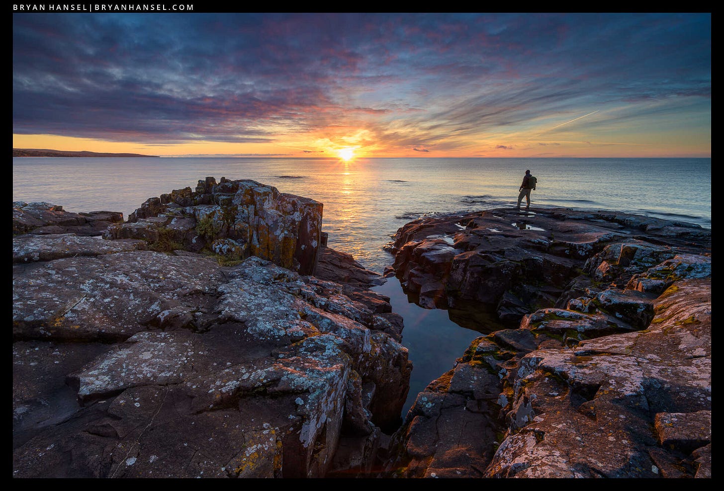 Fiery clouds over a basalt shoreline with a person looking small under the bigness of the scene. 