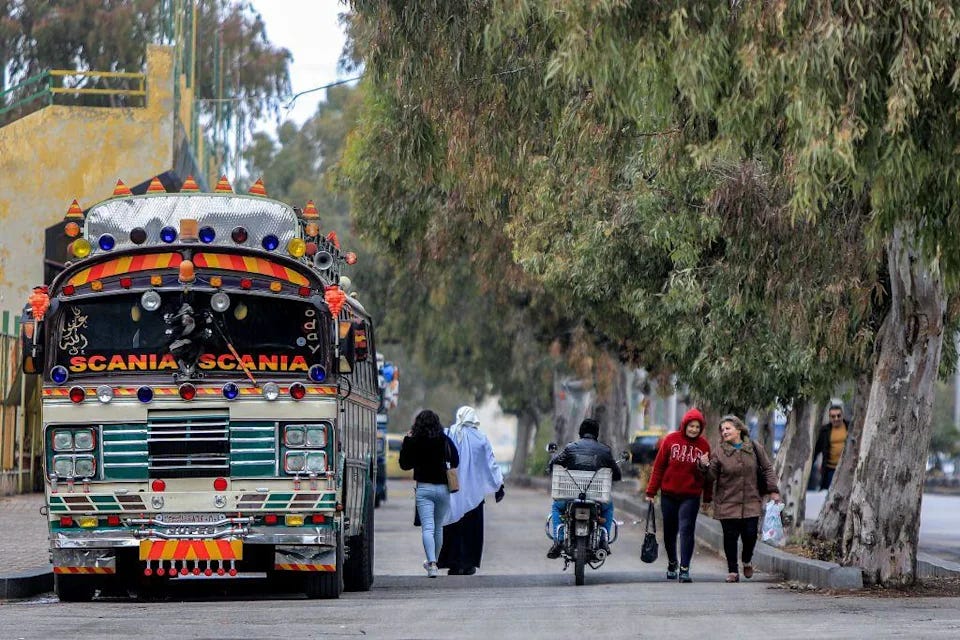 A la izquierda de una carretera, un autobús urbano lleno de farolillos y señas de colores. En la misma vía van caminando por el centro dos mujeres, una de ellas con velo en la cabeza, después una moto y otras dos mujers más. Al lado derecho de la vía hay una acera llena de árboles. A la izquierda de una carretera, un autobús urbano lleno de farolillos y señas de colores. En la misma vía van caminando por el centro dos mujeres, una de ellas con velo en la cabeza, después una moto y otras dos mujers más. Al lado derecho de la vía hay una acera llena de árboles.