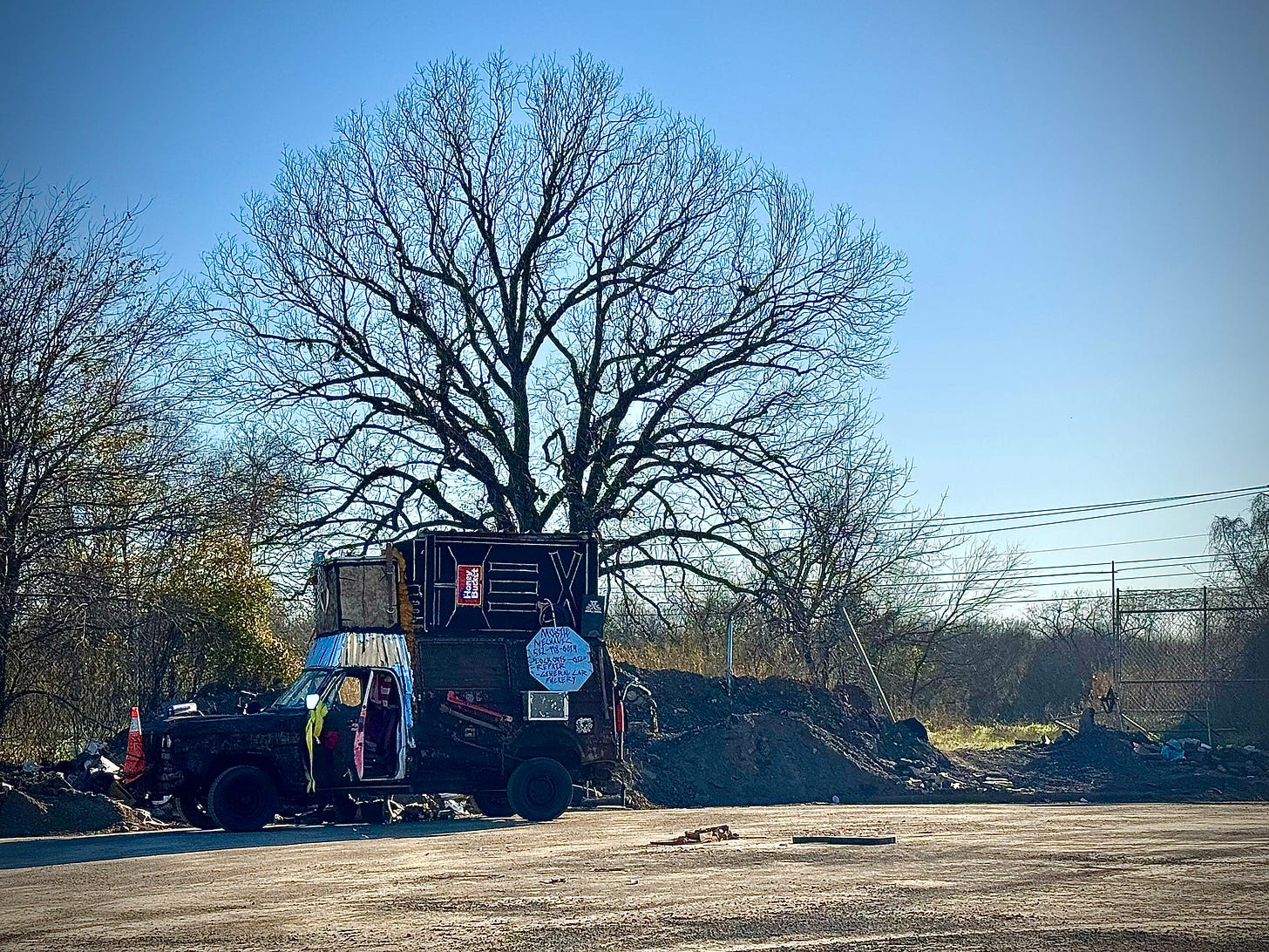 Heavily hand-modified pickup with a Honey Bucket on top and a dude working underneath, next to a pile of asphalt at a cul de sac with a cut chain link fence opening into an expanse of urban woods