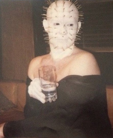 HELLRAISER behind-the-scenes photo of actor Doug Bradley as Pinhead. He's in full white face paint with his titular head full of pins, but he's clearly a work in progress: only one hand painted white, rest of his skin unpainted, loosely draped in a black bathrobe that drapes coyly off his shoulders, and holding a glass of water in his (painted) hand like it's a wineglass. The vibe is Real Housewives of the Hell Dimension