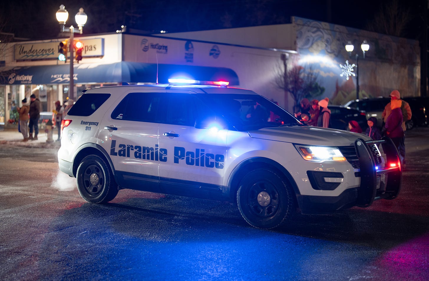A white SUV with "Laramie Police" written on the side and lights on crosses an intersection at night. Warmly bundled pedestrians are visible in the background.