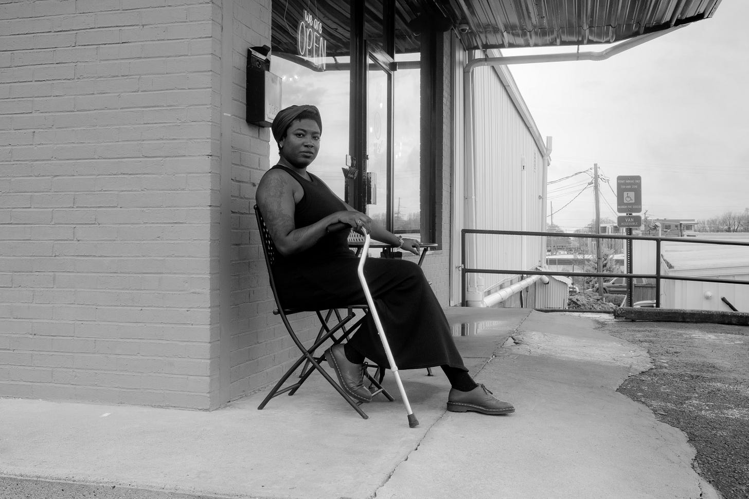 Black and white photo of the author in a black dress seated with her cane in front of a coffeeshop and an accessible parking sign. Black and white photo of the author in a black dress seated with her cane in front of a coffeeshop and an accessible parking sign.