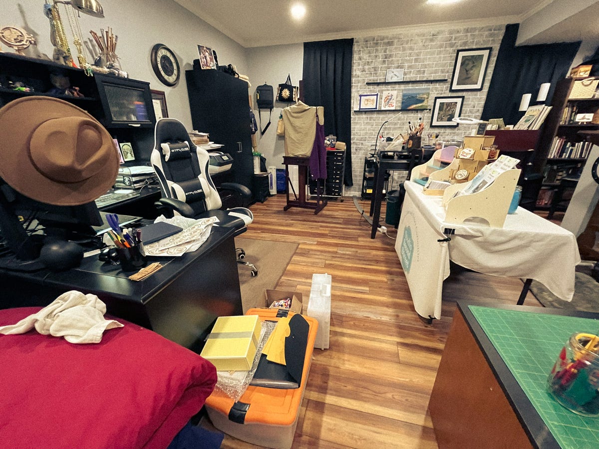 A warmly lit creative studio filled with organized chaos—desks stacked with paper, tools, and zines in progress. A tan felt hat rests on a computer monitor, a white-and-black chair sits at one desk, and a display table holds zine racks and art supplies under soft gray walls and brick accents.