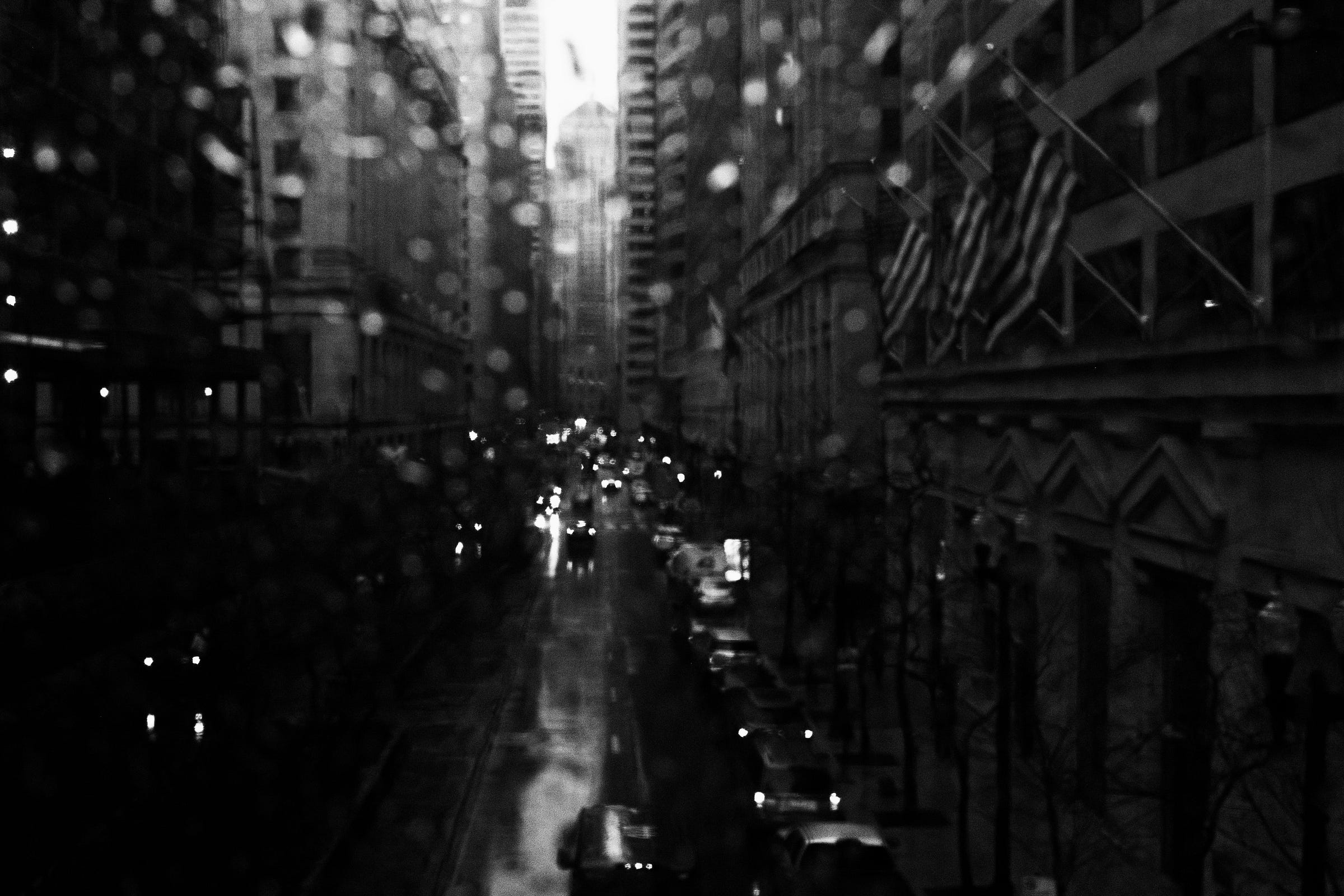Rain-speckled window overlooking a wet city street lined with towering buildings, American flags, and glowing headlights reflecting off the pavement. Rain-speckled window overlooking a wet city street lined with towering buildings, American flags, and glowing headlights reflecting off the pavement.