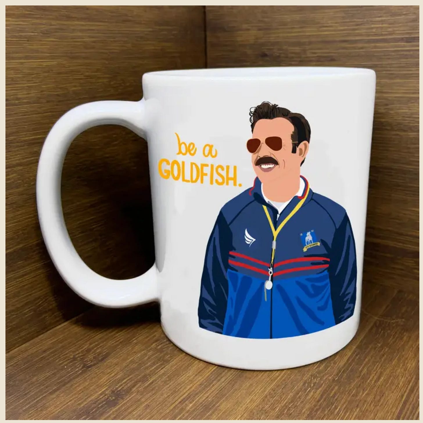 Mug with Ted Lasso on it and in gold letters the phrase "be a goldfish"