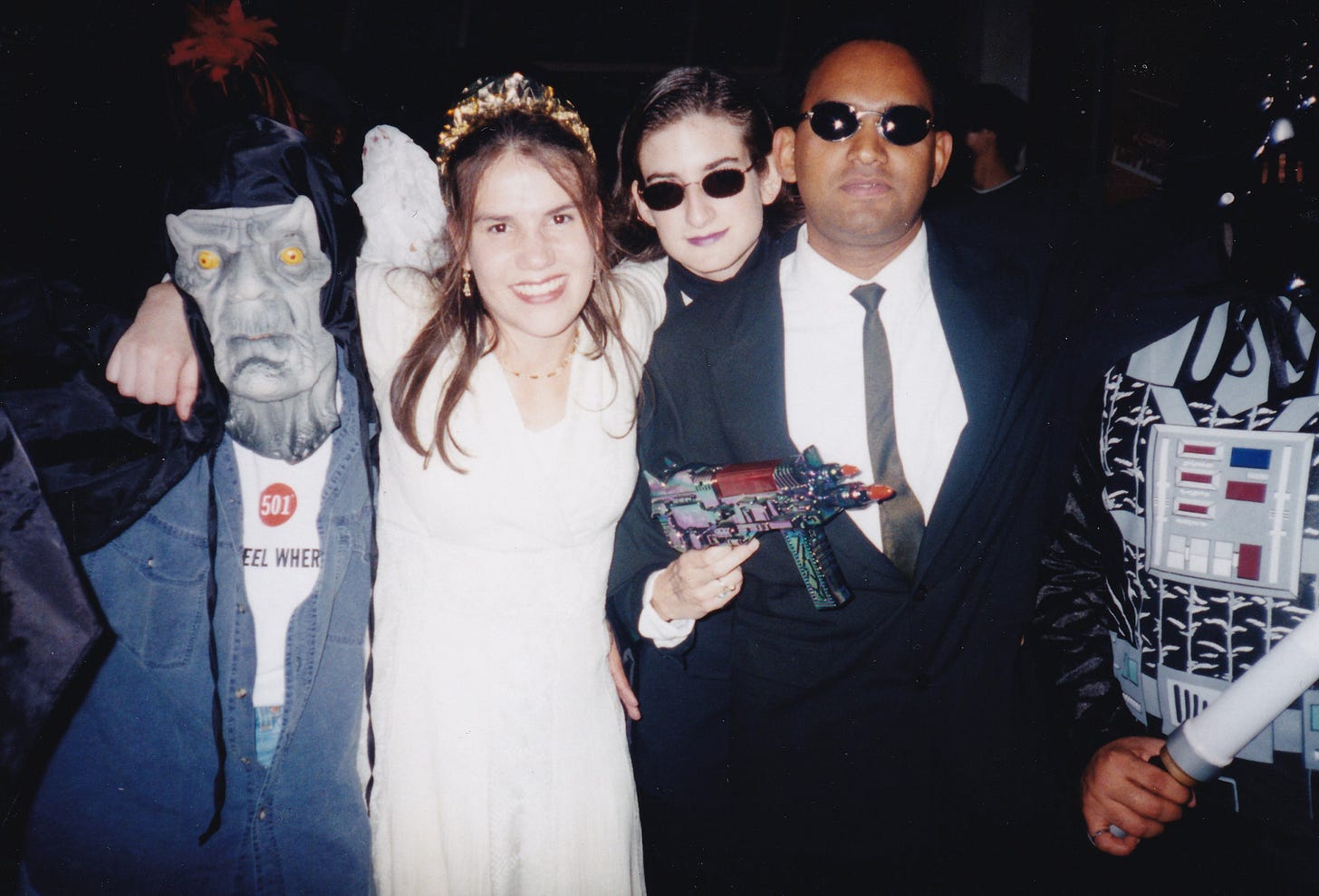Inside a bar at night, five adults stand together. A man in an emperor’s mask, a woman dressed like an angel, a woman and man dressed like the characters from Men in Black, and a man dressed like Darth Vader. 
