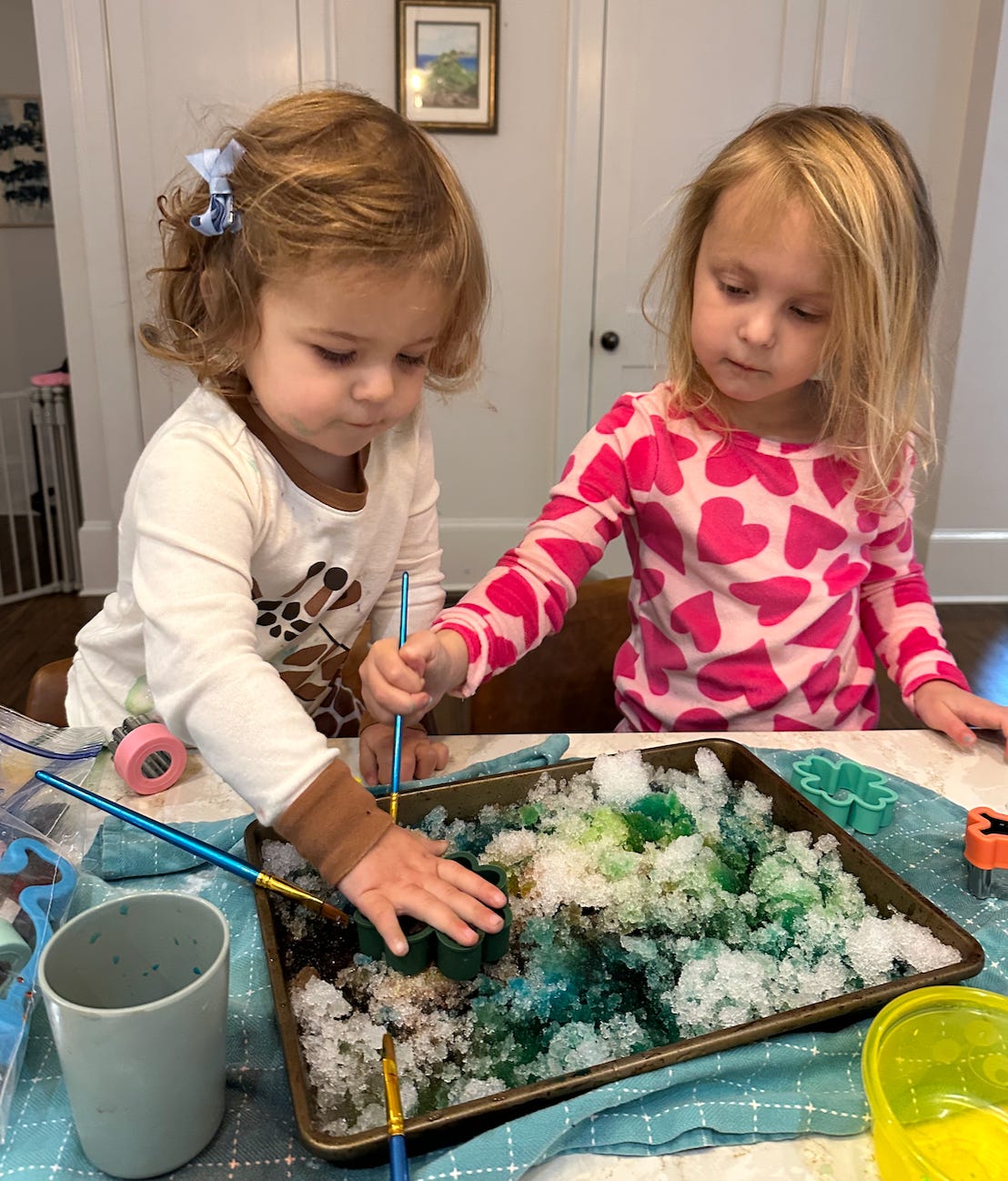 Two kids are engrossed in an activity table, coloring a tray filled with white crystals with paintbrushes and stamping out shapes with cookie cutters.