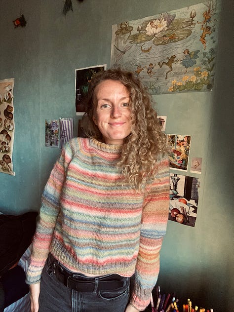 JB wearing a pastel coloured striped jumper that she knitted. She is standing in said jumper looking very pleased, with her thumbs up and a smile on her face.