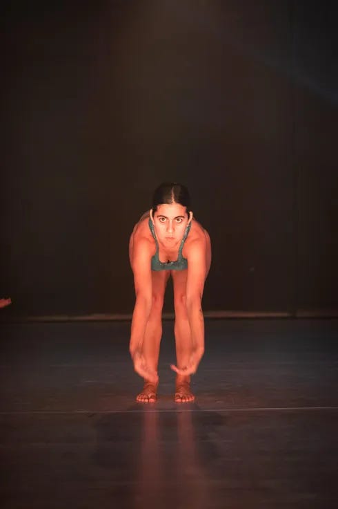 Vue frontale d’une danseuse pieds nus en justaucorps turquoise et argenté, penchée profondément vers l’avant, les bras tendus et les mains proches du sol. Éclairée par une lumière chaude sur une scène sombre, elle regarde intensément droit devant elle. Vue frontale d’une danseuse pieds nus en justaucorps turquoise et argenté, penchée profondément vers l’avant, les bras tendus et les mains proches du sol. Éclairée par une lumière chaude sur une scène sombre, elle regarde intensément droit devant elle.