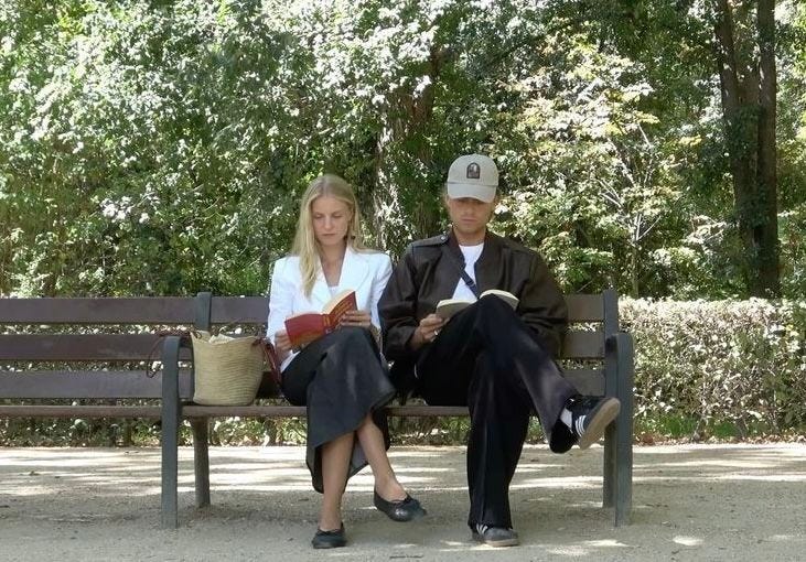 This may contain: a man and woman sitting on a bench in front of some trees, reading books