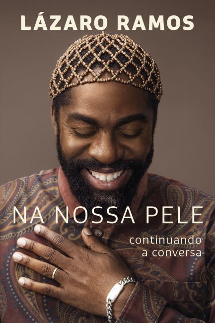 #descrevemospravocê  Capa do livro Na Nossa Pele: continuando a conversa, de Lázaro Ramos. O título principal aparece em letras brancas, centralizado na parte inferior da imagem, enquanto o nome do autor está em destaque na parte superior, também em letras brancas e em caixa alta. Abaixo do título, à direita, aparece a frase complementar “continuando a conversa”, em letras menores.  A imagem de fundo é uma fotografia de Lázaro Ramos. Ele está com os olhos fechados e sorrindo suavemente, com expressão de serenidade e afeto. Sua mão direita está apoiada sobre o peito, como em um gesto de respeito ou gratidão. Lázaro usa uma camisa estampada com desenhos em tons terrosos e arabescos, além de uma pulseira metálica no pulso direito e uma aliança no dedo anelar da mão esquerda. Na cabeça, ele usa um acessório trançado de miçangas em tons de dourado e marrom, que envolve os cabelos curtos. O fundo da capa é liso, em tom marrom claro, o que ajuda a destacar a figura do autor.  A composição visual transmite acolhimento, reflexão e identidade, elementos que dialogam com o tema do livro.