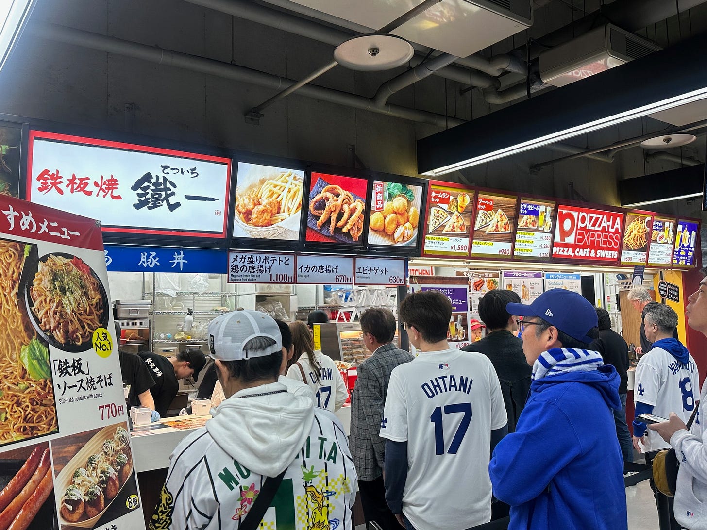 A photo of fans, mostly in Dodger jerseys, waiting in line to purchase concessions--including shrimp nuggets, fried squid, and stir fried noodles with sauce