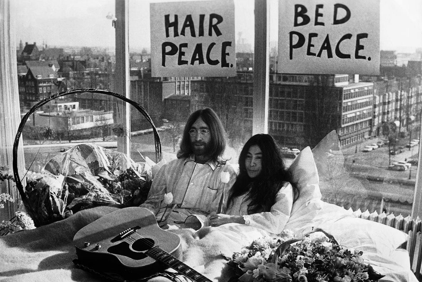 A classic black and white photograph of John Lennon and Yoko Ono sitting up in bed during their 'Bed-In for Peace,' with hand-drawn signs behind them reading 'HAIR PEACE' and 'BED PEACE.'