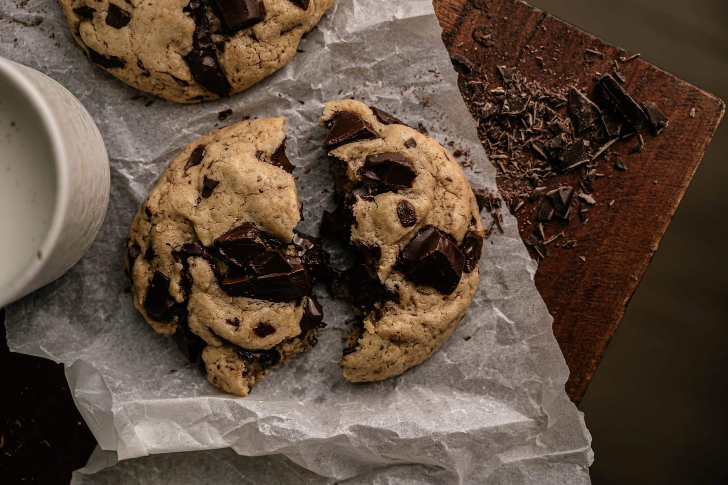 a ripped in half fresh small batch chocolate chip cookie on a table of chocolate crumbs and a glass of milk. 