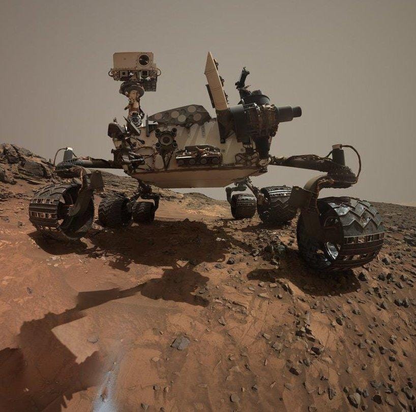 A low-angle color photo of the Curiosity rover, its six wheels resting on the rocky Martian surface, with a grey/brown sky behind it A low-angle color photo of the Curiosity rover, its six wheels resting on the rocky Martian surface, with a grey/brown sky behind it