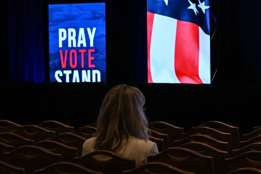 A woman wearing a tan jacket is seen from behind sitting in a chair and facing a stage where screens display an American flag and a sign reading “Pray Vote Stand.”