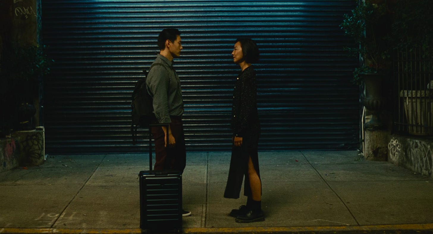 Nora e Hae Sung em pé frente a frente na calçada à noite, com uma porta metálica fechada ao fundo. Ele segura uma mala, sugerindo um momento de despedida na última noite em Nova York.