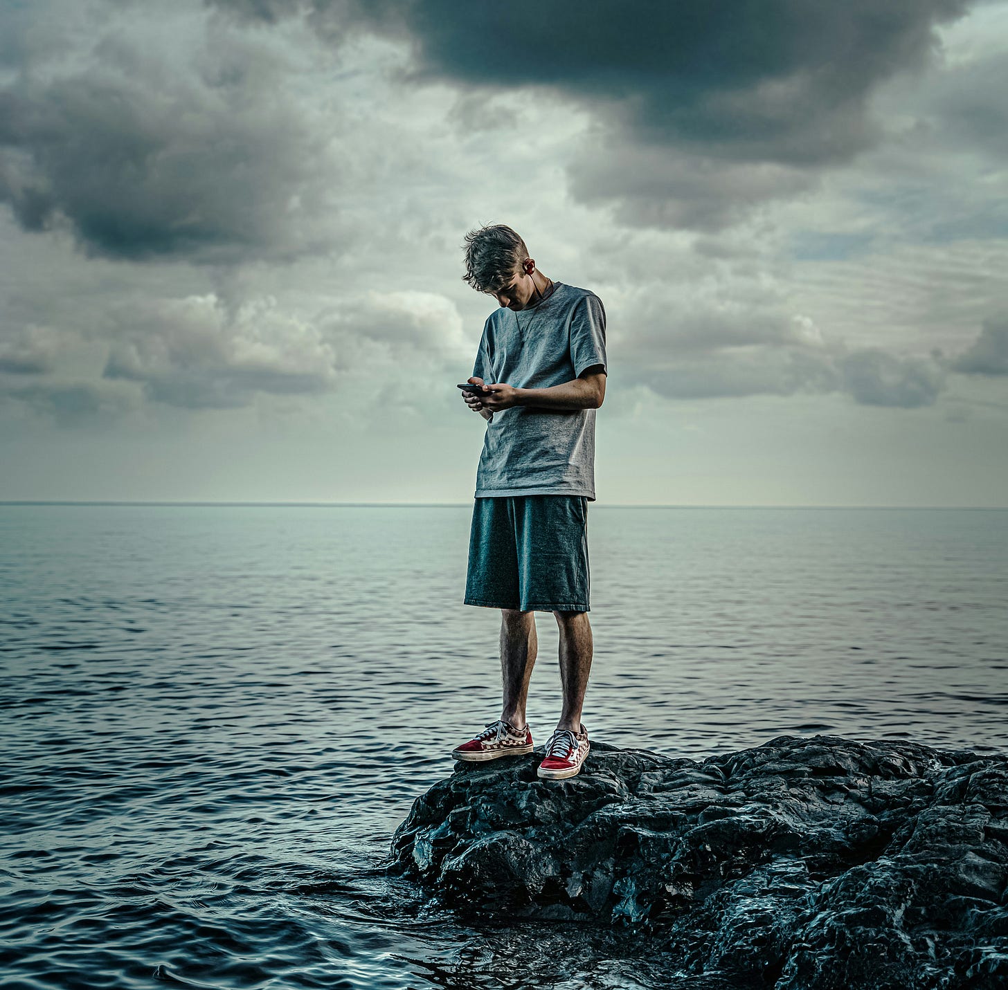 A man in black shorts, a white t-shirt and red sneakers standing on the edge of a rock pool next to a vast and still sea. His head is down and he’s looking at his phone, which is in his hands in front of his chest.