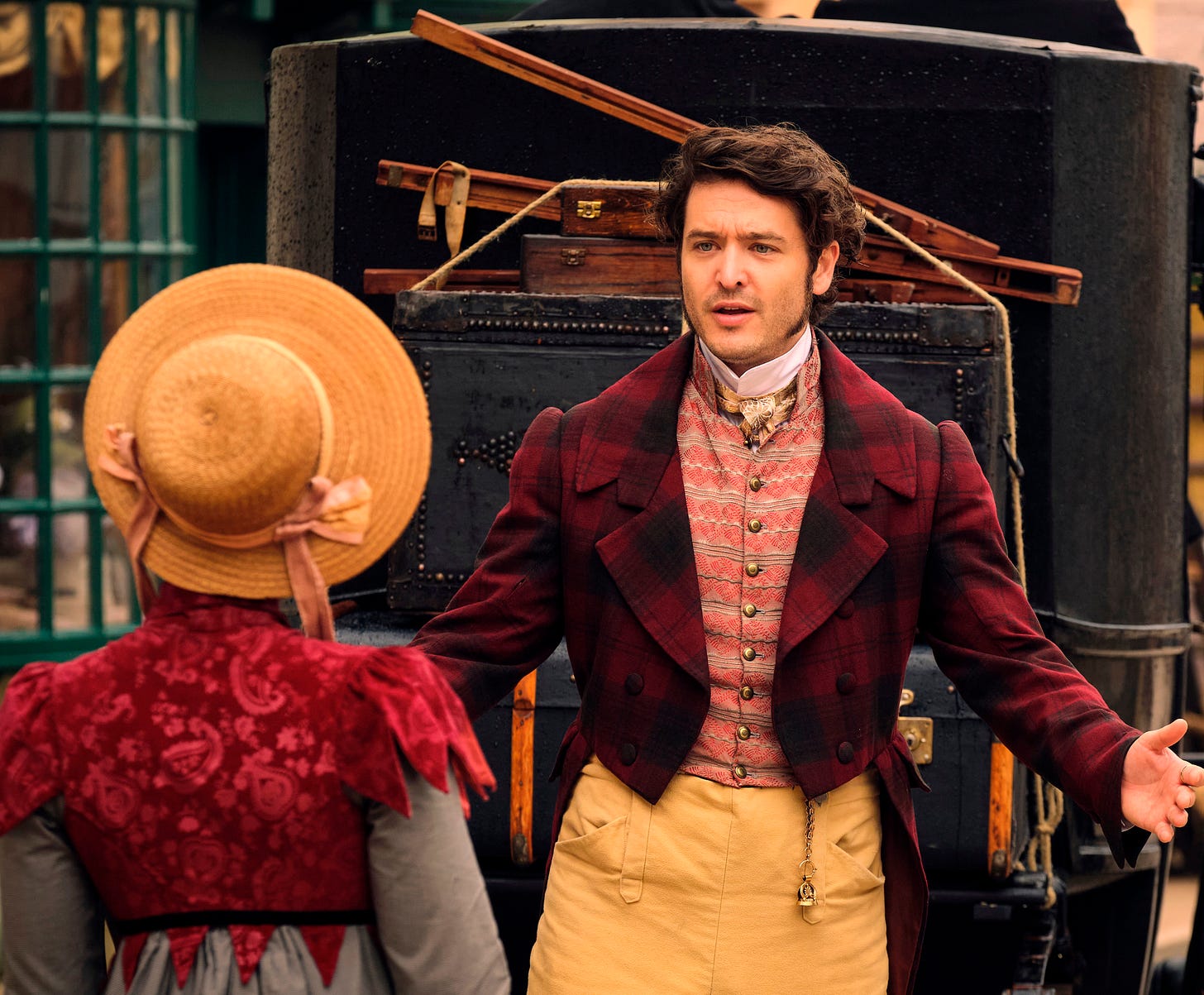 A young man in Regency fashion holds his arms out and talks with a young woman in a red Regency-style jacket and gown, her back to the camera
