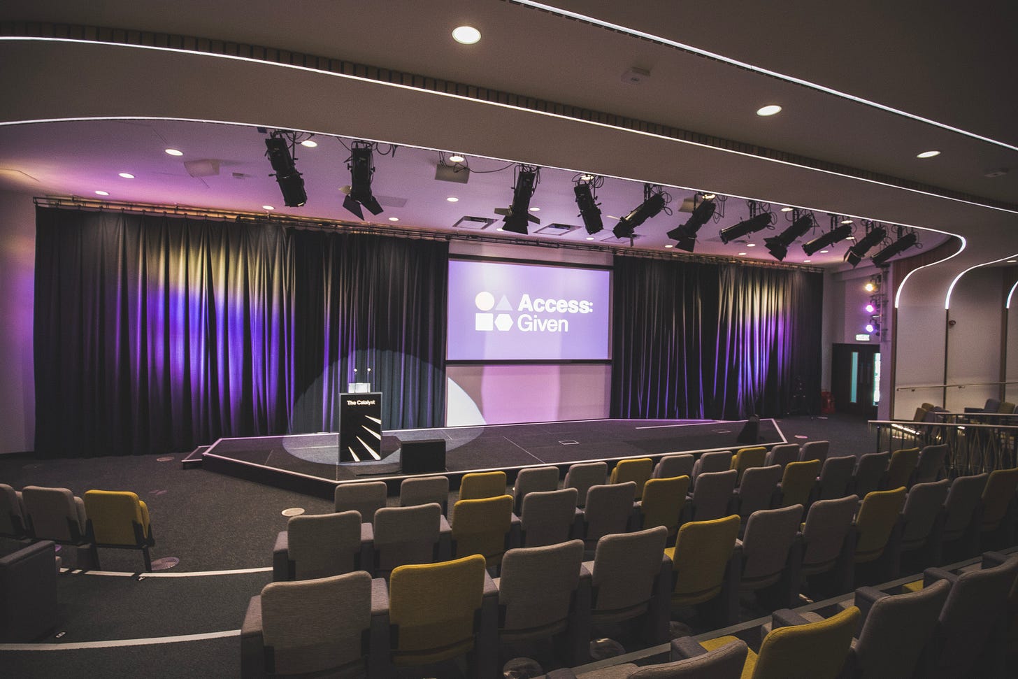 An empty auditorium with rows of gray and yellow chairs facing a stage. On stage, a podium labeled "The Catalyst" is positioned to the left. Behind it, a large screen displays the logo and text “Access: Given” with simple geometric icons. The stage is lit with purple and yellow spotlights against dark curtains, and multiple ceiling lights and rigged stage lights are visible above.