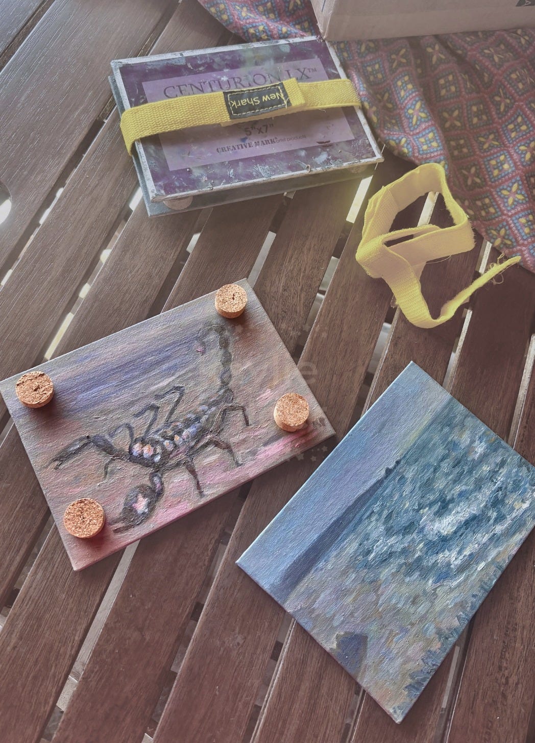 small oceanside paintings with cork slices in each corner, and two painting panels sandwiched together with velcro straps on wood slat table