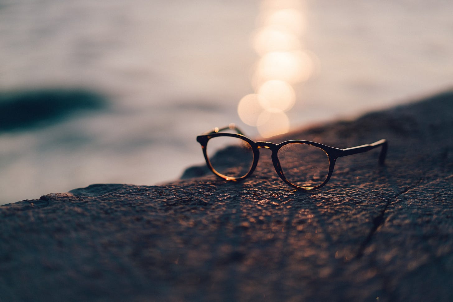 Muretto di pietra con un paio di occhiali aperti appoggiatici sopra. Lo sfondo grigio è sfumato e sembra una distasa marina, illuminata dalla luce del sole al tramonto. Muretto di pietra con un paio di occhiali aperti appoggiatici sopra. Lo sfondo grigio è sfumato e sembra una distasa marina, illuminata dalla luce del sole al tramonto.