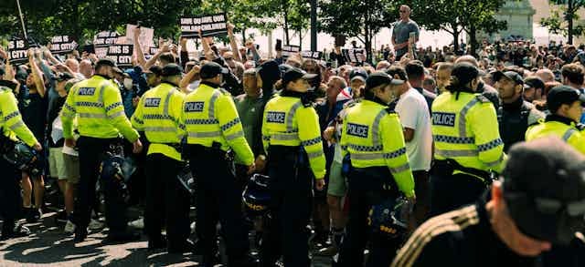 A line of police in hi-vis jackets facing a large crowd of mostly white men, holding signs that read 'save our children'. A line of police in hi-vis jackets facing a large crowd of mostly white men, holding signs that read 'save our children'.