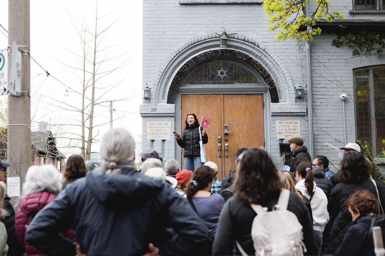 Rebecca Arshawsky holding a pink sign leading a group of 100 people on a Jane's Walk in front of a synagogue.