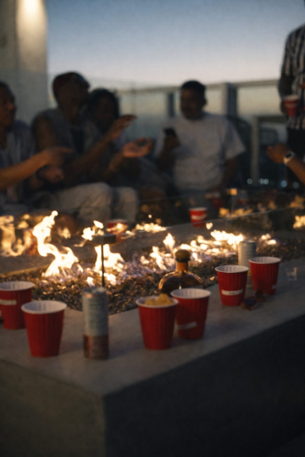 Rooftop fire pit glowing at dusk with flames in the foreground, red cups along the ledge, and indistinct figures seated beyond the fire against an evening sky.