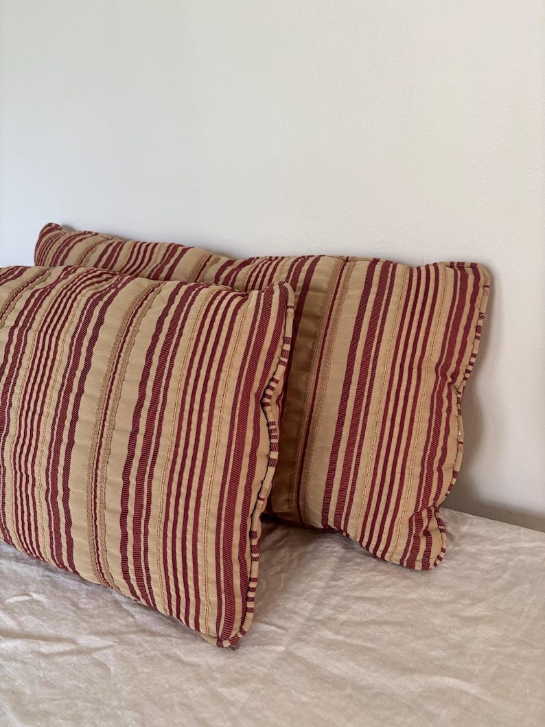May include: Two rectangular throw pillows featuring a vertical striped pattern in deep red and beige. The fabric has a textured appearance with piped edges along the perimeter, creating a structured look for these soft, stuffed decorative cushions.