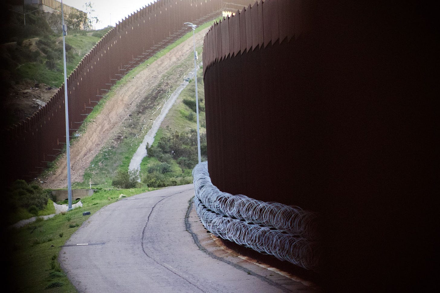 Two layers of tall, brown bollard walls cut through a hilly landscape. Rows of concertina wire is visible at the base of the wall on the right. Two layers of tall, brown bollard walls cut through a hilly landscape. Rows of concertina wire is visible at the base of the wall on the right.