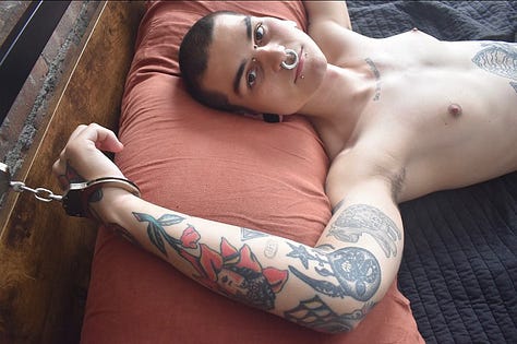 A collage of photos of Rae in various forms of bondage: ranging from chained to a car hoist, to rope and rope suspension as well as cuffs and leather bound to a bed, or their ankles. Rae has many tattoos on their arms, legs and stomach. They have a shaved head and brown eyes. They have a large septum ring, a bridge piercing, and stretched earlobes.