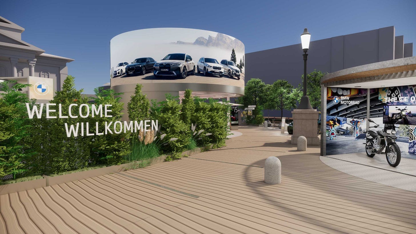 Una imagen 3D de un moderno centro comercial al aire libre. El elemento principal es un gran edificio cilíndrico con un anuncio envolvente de coches. El edificio es blanco con un cielo azul y nubes reflejadas en él. El anuncio muestra varios coches sobre un fondo blanco. El primer plano tiene una pasarela de madera con un cartel de “Bienvenido” en inglés y alemán. También hay varias tiendas y restaurantes en el fondo. La escena está ambientada durante el día con un cielo azul claro.