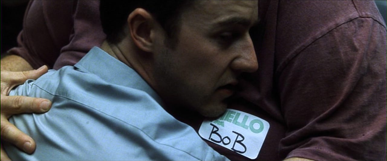 A screenshot from Fight Club, in which Edward Norton's character (white, brown hair, blue business shirt) embraces Bob (not in scene, red shirt, heavy) and looks really sad