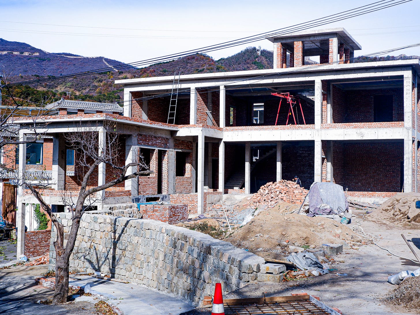Raw concrete pillars, exposed brick, and dirt mounds mark the construction of new guesthouses, against an historical backdrop.