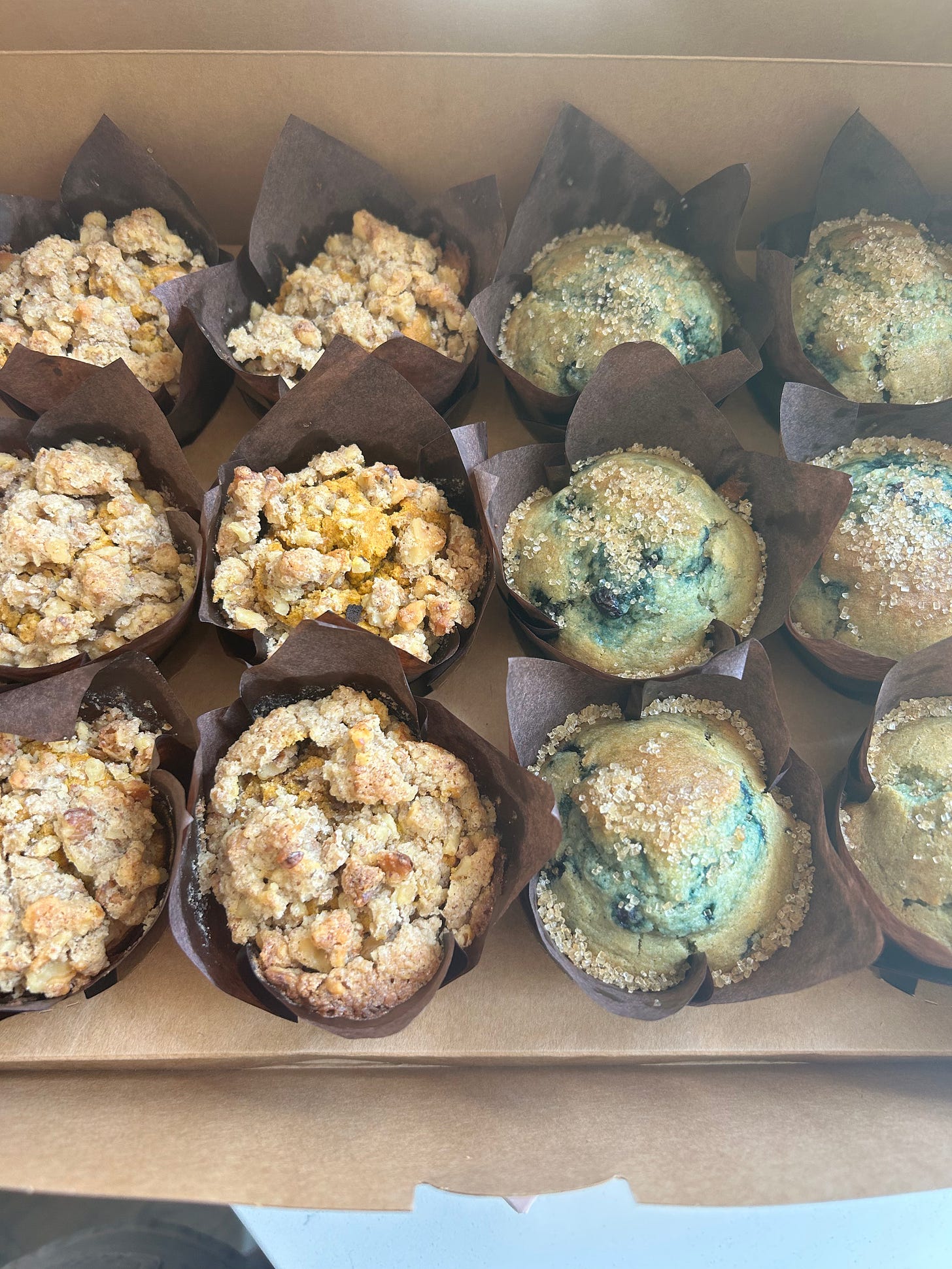 A bakery box of gluten-free muffins in brown paper liners, crumb-topped muffins on the left and blueberry muffins on the right.